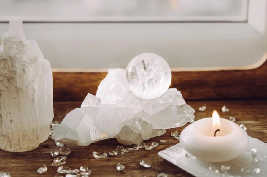 A serene arrangement featuring white selenite healing crystals on a wooden surface near a window. The display includes a selenite crystal sphere, clusters of raw selenite crystals, scattered small stones, and a white candle burning in the foreground.