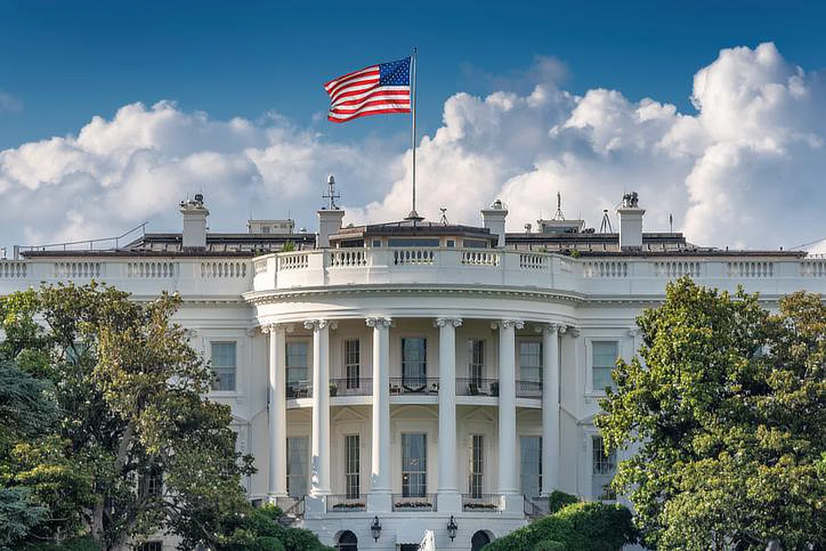 The White House with the U.S. flag flying on top, framed by trees in the foreground, set against a partly cloudy sky.