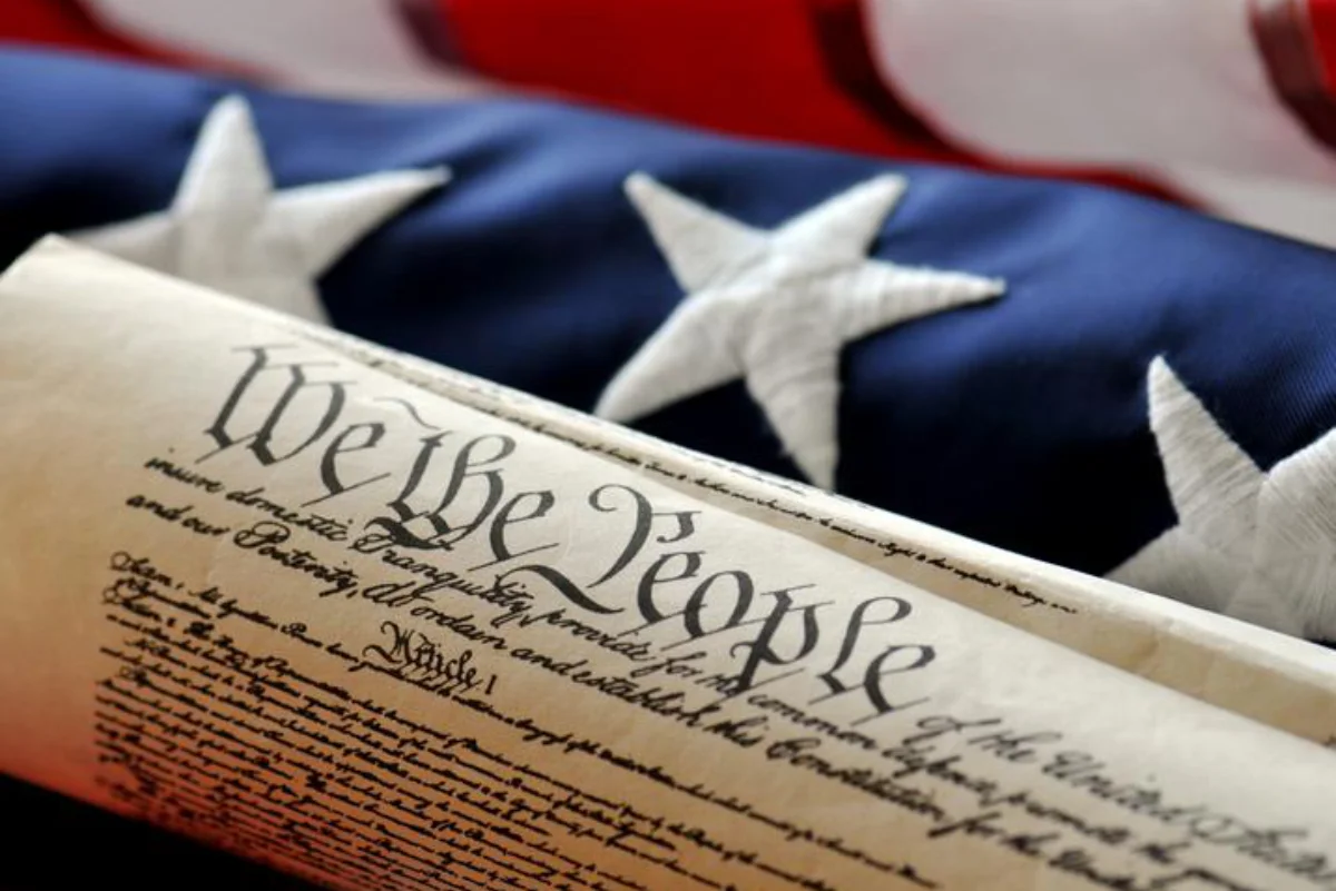 Rolled-up United States Constitution document with the words 'We the People' prominently displayed, placed in front of an American flag with visible white stars on a blue background.