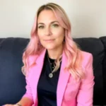 Priscilla Daphney sitting on a couch, wearing a pink blazer and black top, looking confidently at the camera.