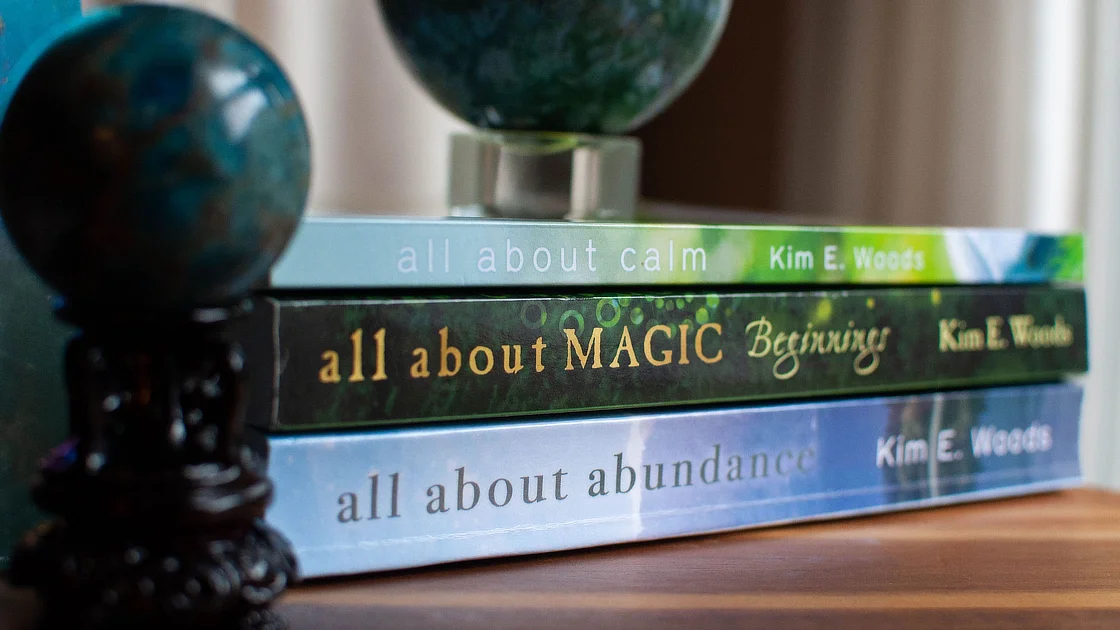 A close-up of three stacked books by Kim Woods: "All About Calm," "All About Magic," and "All About Abundance," with a decorative globe nearby.