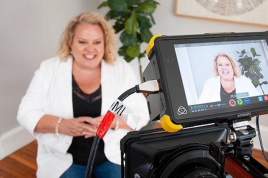 Kim Woods sits in front of a camera during a video recording session, smiling and wearing a white blazer. The camera screen shows her image as she speaks, with a plant in the background.