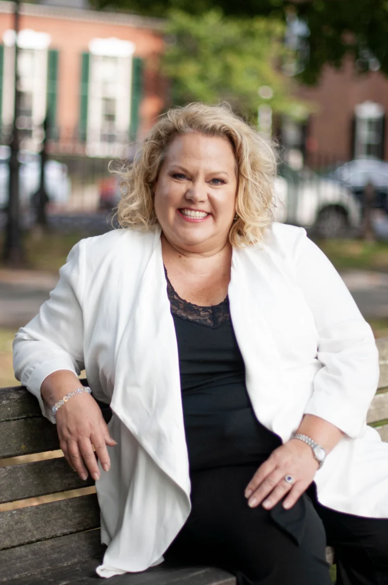 Kim Woods, smiling and seated on a bench outdoors, wearing a white jacket over a black top, with a blurred background of trees and buildings.