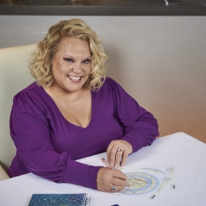 Kim Woods smiling while drawing on an astrological chart at a table, wearing a purple top with a notebook beside her.