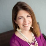 Bridgette Barr, a woman with long brown hair smiling brightly, wearing a vibrant purple blouse and layered beaded necklaces, seated in a modern indoor space.