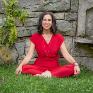 Carla Savetsky, a smiling woman sitting cross-legged on the grass, wearing a vibrant red jumpsuit and a pendant necklace, with a serene stone wall and greenery in the background.