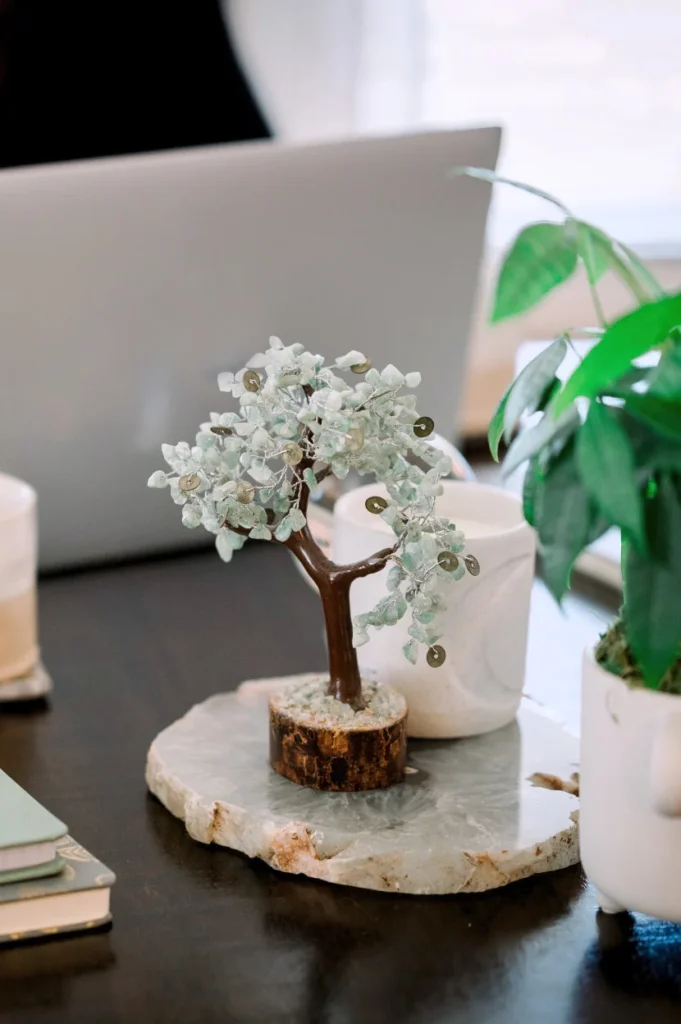 A decorative gemstone money tree on a polished stone base, displayed on a desk with notebooks, plants, and a laptop in the background.