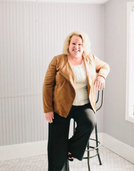 Kim Woods leaning casually on a tall chair, wearing a tan suede jacket, white blouse, and black pants, in a bright room with white paneled walls and natural light streaming through a window.