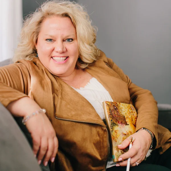 Kim Woods sitting on a couch holding a colorful journal, dressed casually in a tan jacket and white blouse in a cozy indoor setting.