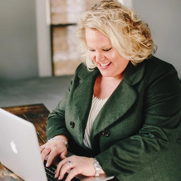 Kim Woods with a cheerful expression sitting at a wooden desk, typing on a laptop in a bright indoor setting.