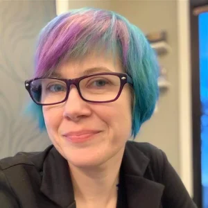 Miranda Burt, a woman with vibrant purple and teal short hair, wearing glasses and a black jacket, smiling warmly in an indoor setting.