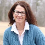 Susan Lynch, a smiling woman with glasses and shoulder-length brown hair, wearing a light blue cable-knit sweater over a white blouse, sitting outdoors with a natural, blurred background.