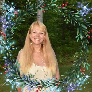 Lisa Nichols, Kim Woods' client, standing outdoors, surrounded by a decorative wreath of green leaves, red berries, and sparkling accents. A forested background is visible behind her, adding to the natural, festive setting.