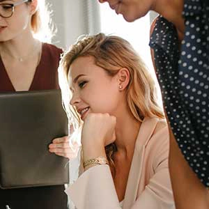 A young woman in a professional setting, smiling thoughtfully as she collaborates with colleagues who are partially visible.