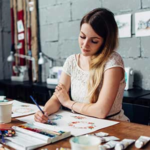 A woman artist painting at a desk, surrounded by art supplies in a studio with gray brick walls.