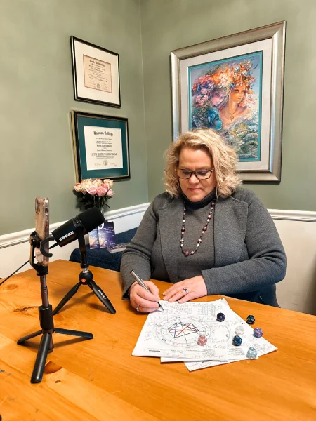 Kim Woods  writing on papers at a desk, with a microphone and a variety of crystals in front of her.