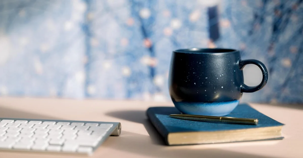 A dark blue speckled mug on a notebook with a gold pen, next to a white keyboard, against a blurred blue background.