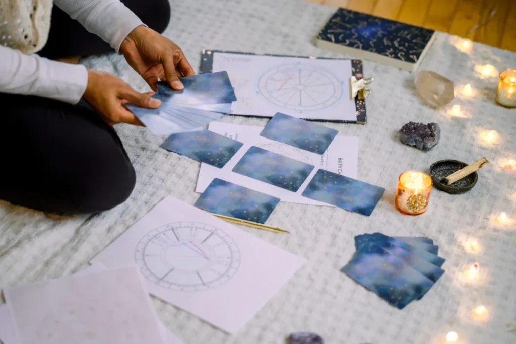 A person sitting on a soft blanket, laying out tarot or oracle cards in a spread, surrounded by astrology charts, candles, crystals, and a smudging dish, creating a mystical and intuitive atmosphere.