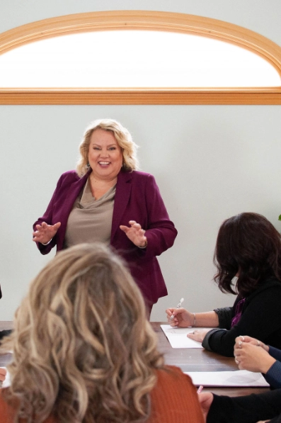 Kim Woods in a purple blazer stands at the head of a table, speaking animatedly to a group of women who are taking notes. The setting is a bright room with an arched window.