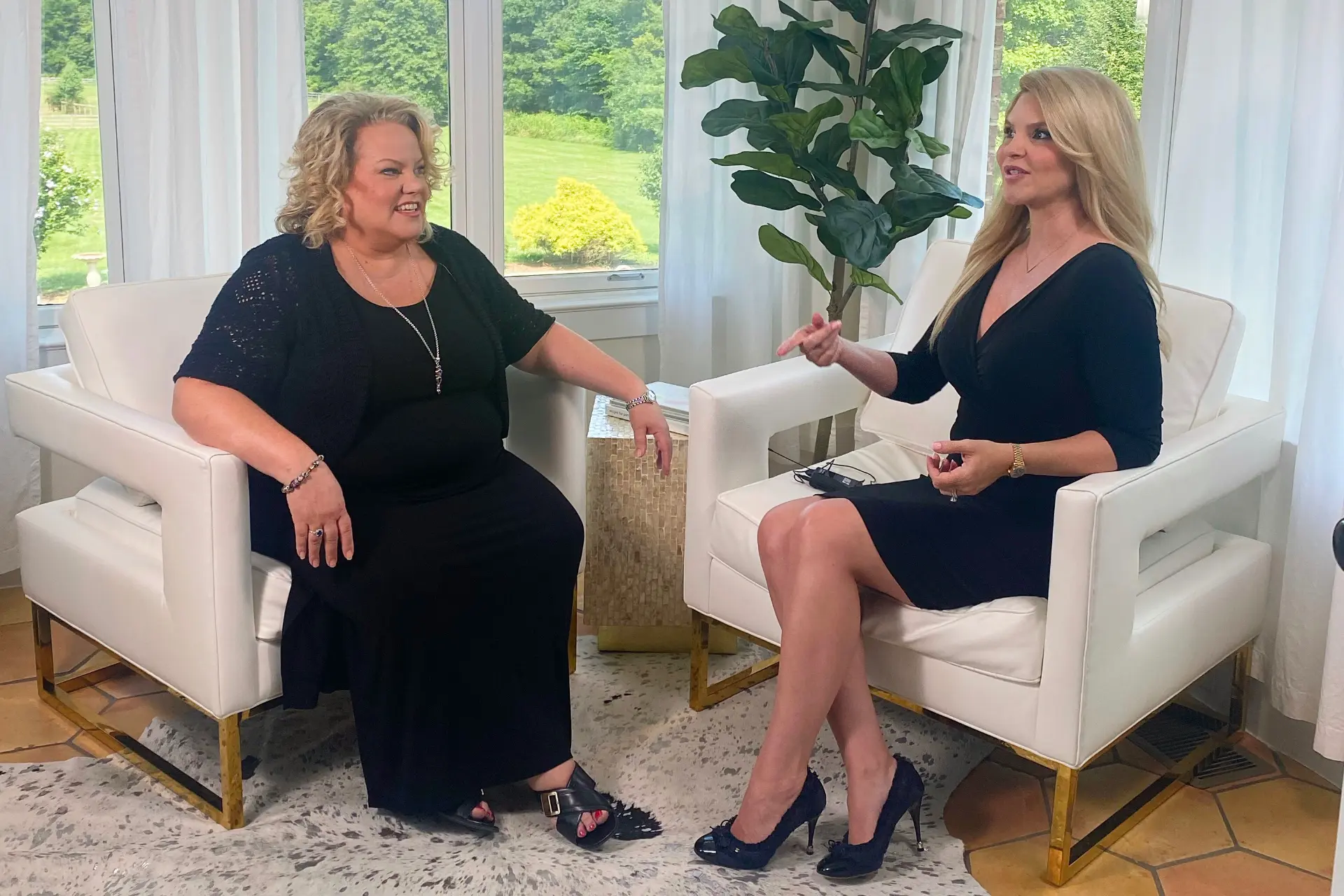 Kim Woods and Brielle Cotterman sit in white armchairs, engaged in conversation. The blonde woman on the left listens with a smile, while the woman on the right gestures as she speaks. The setting is a bright, elegant room with large windows and greenery outside.