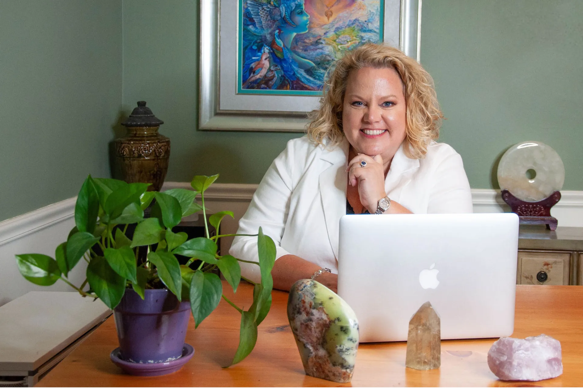 Kim Woods sits at a desk with a MacBook, smiling. Crystals and a potted plant decorate the desk, with a mystical painting on the green wall behind her.