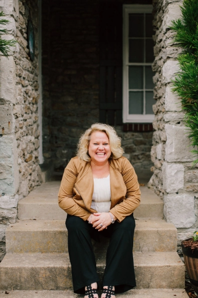 Kim Woods in a tan jacket and black pants sits on stone steps, laughing warmly. She is framed by a rustic stone doorway with greenery on either side.