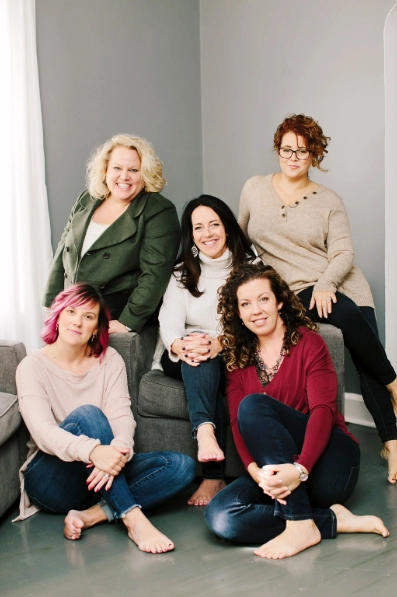 A group of Kim Woods' clients five women sits together in a cozy, light-filled room, dressed in casual sweaters and jeans. They smile warmly, exuding a sense of connection and camaraderie.