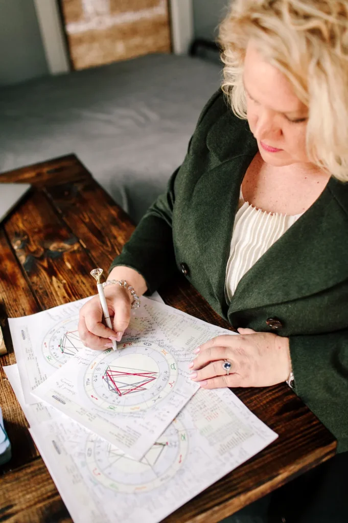 Kim Woods in a dark green coat studies astrology charts at a wooden table, writing notes with a pen topped with a crystal.