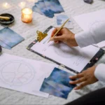 A person fills out a birth chart on a clipboard, surrounded by astrology charts, tarot cards, crystals, and candles. The setting is a cozy, mystical space, perfect for intuitive and astrological insights.