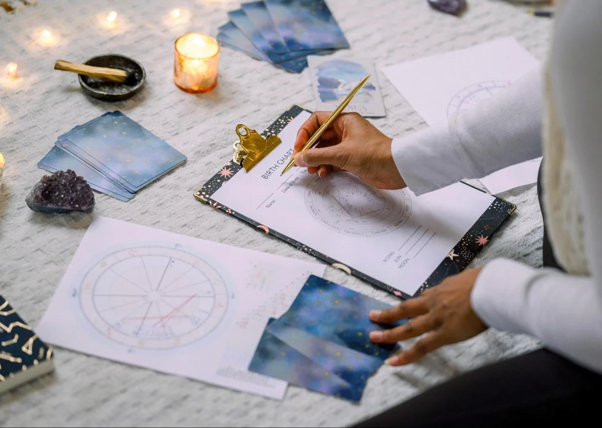 A person fills out a birth chart on a clipboard, surrounded by astrology charts, tarot cards, crystals, and candles. The setting is a cozy, mystical space, perfect for intuitive and astrological insights.