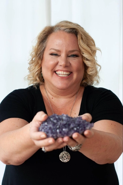 Kim Woods wearing a black top and silver jewelry, smiles brightly while holding a large cluster of purple amethyst crystals in both hands. She stands in front of a simple white backdrop, exuding joy and warmth.