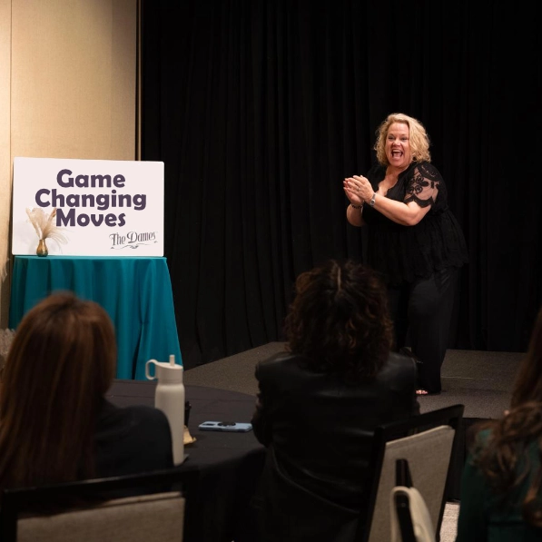 Kim Woods in a black lace top speaks energetically on stage, clapping her hands. A sign beside her reads "Game Changing Moves." The audience watches attentively in a professional event setting.