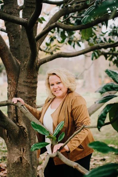 Kim Woods in a tan suede jacket and white top poses outdoors among the branches of a large tree. She leans on a branch with one hand and smiles warmly at the camera. The background features a park-like setting with trees and fallen leaves.