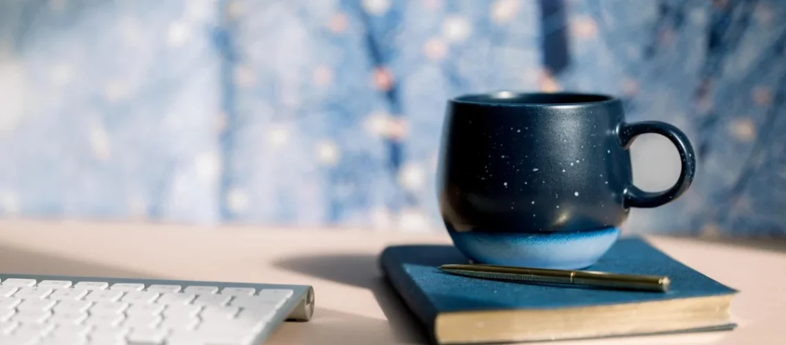 A dark blue speckled mug on a notebook with a gold pen, next to a white keyboard, against a blurred blue background.