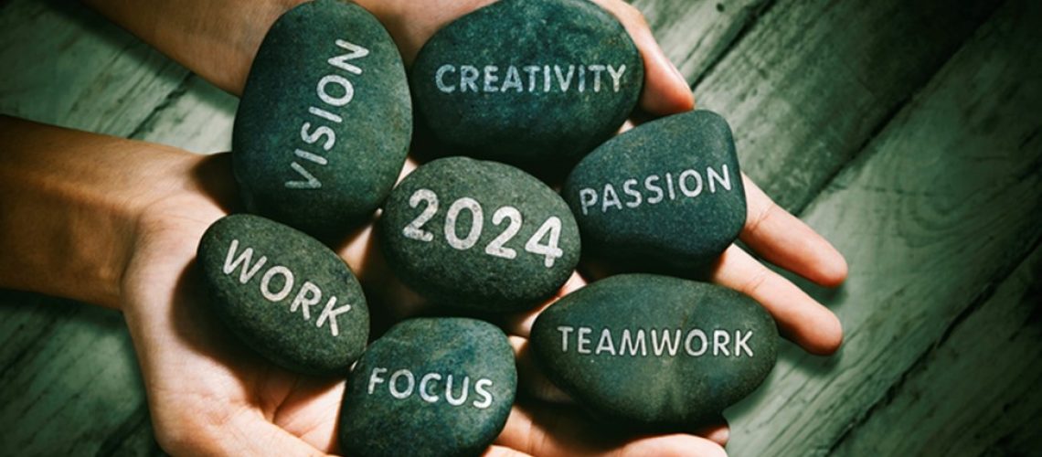 Two hands holding several smooth, black stones. Each stone has a word engraved on it, including 'VISION,' 'CREATIVITY,' 'PASSION,' 'WORK,' 'FOCUS,' 'TEAMWORK,' and '2024.' The stones symbolize motivational values and goals for the year 2024.