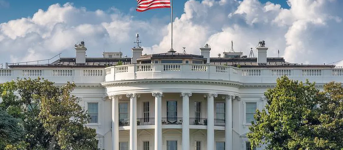 The White House with the U.S. flag flying on top, framed by trees in the foreground, set against a partly cloudy sky.