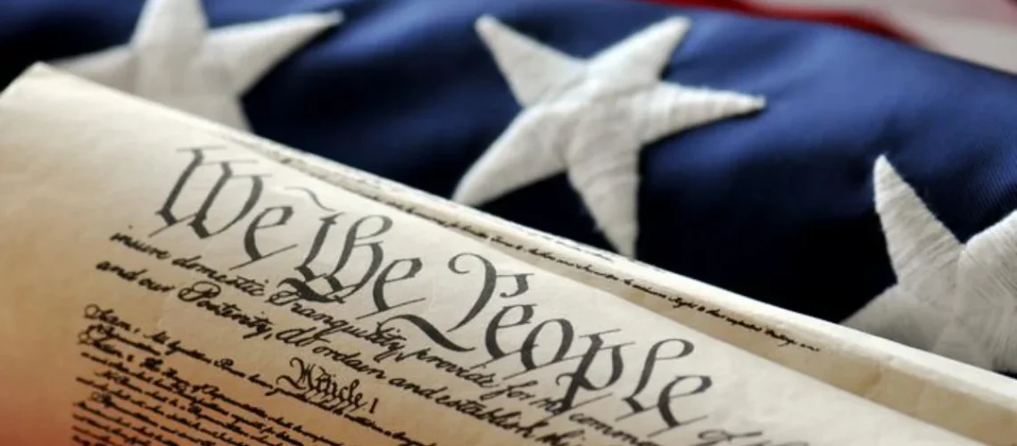 Rolled-up United States Constitution document with the words 'We the People' prominently displayed, placed in front of an American flag with visible white stars on a blue background.